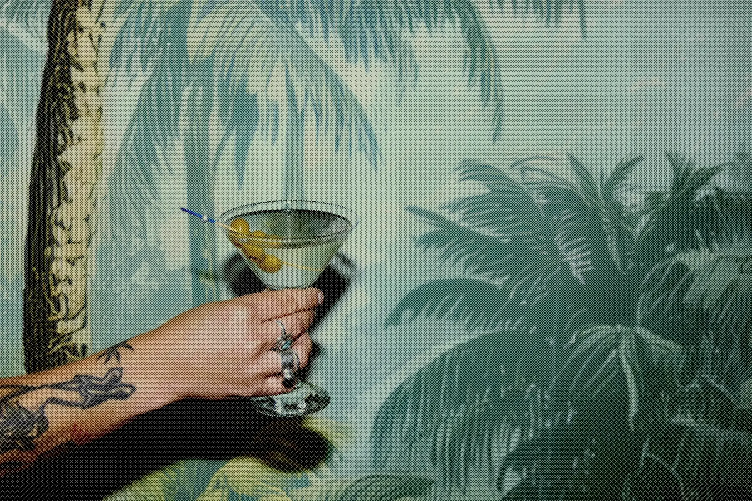A martini with olives held out against the full sweep of the palm-tree wallpaper.